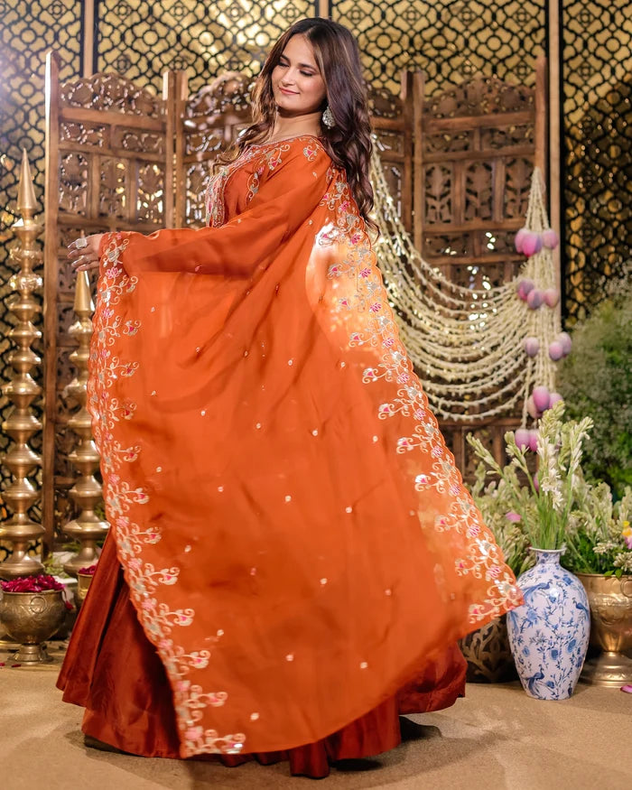 Woman in an orange saree with intricate patterns standing in a decorated indoor setting.