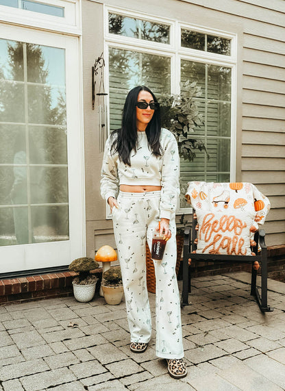 A woman stands on a front porch wearing a matching light gray and white fall-themed lounge set with a subtle pumpkin and leaf pattern. She is holding a coffee cup next to a chair with a 'Hello Fall' blanket.
