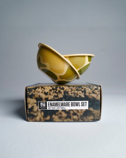 2 camo-patterned enamel bowls sitting intricately balanced on top of box, against studio background