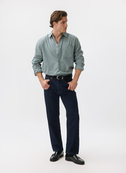 A man models the Percival Clancy Striped Long Sleeve Shirt in blue, styled with dark denim jeans and black loafers.