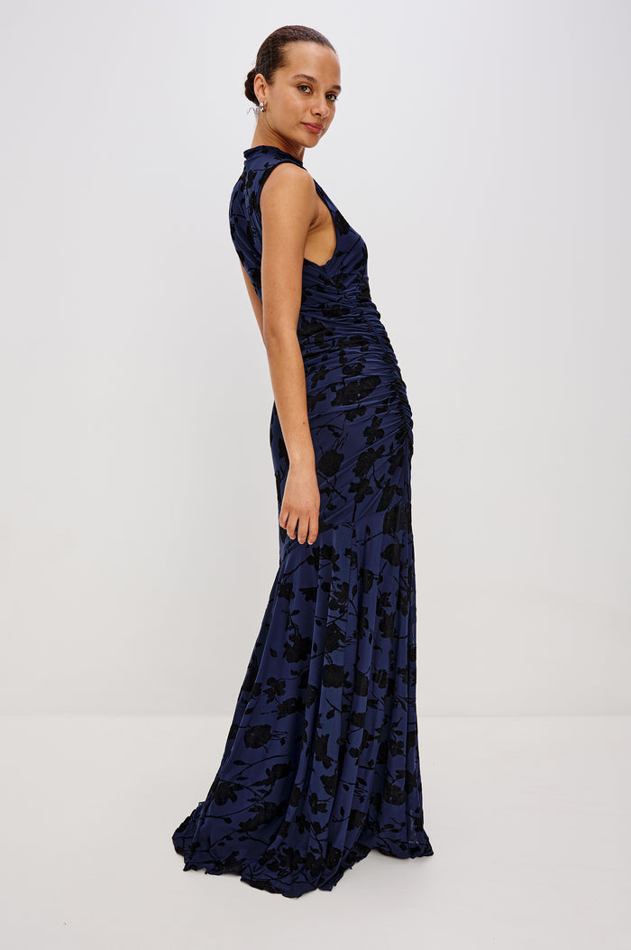 Cosmos Maxi-Length Dress with a Cowl Neck, Sleeveless, Form Fitting with a Ruched Side Seam Detail and an A-line Hem in Navy Tossed Roses Colorway. Full Body Side View Shown on Model.