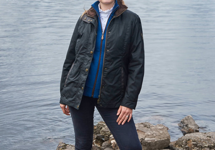 Waist up view of a female model wearing a Dubarry Mountrath Waxed jacket, black jacket with full zip and buttons, brown edged pockets, inner collar and brown inside collar