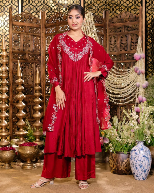 Woman in a red traditional outfit with intricate patterns, standing in a decorative setting with plants and vases.