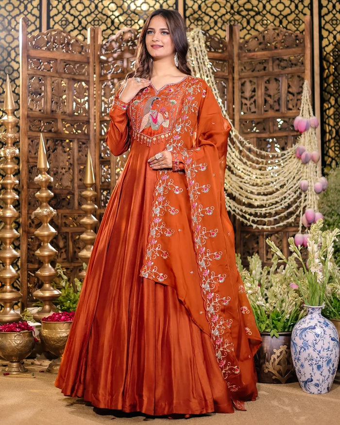 Woman in an orange embroidered traditional outfit standing in a decorative setting with plants and vases.